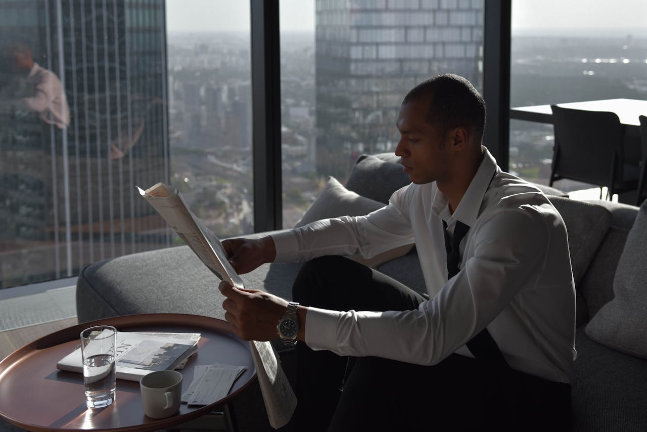 A businessman reads a newspaper by the window with a city view from a high-rise apartment.